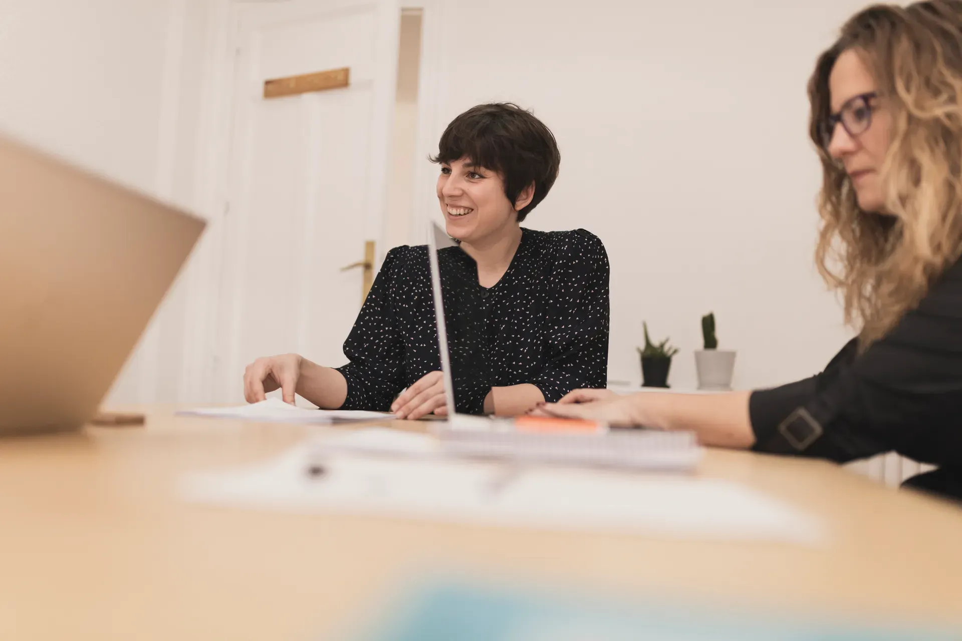 Zwei Frauen arbeiten lächelnd mit Laptops an einem Schreibtisch zusammen